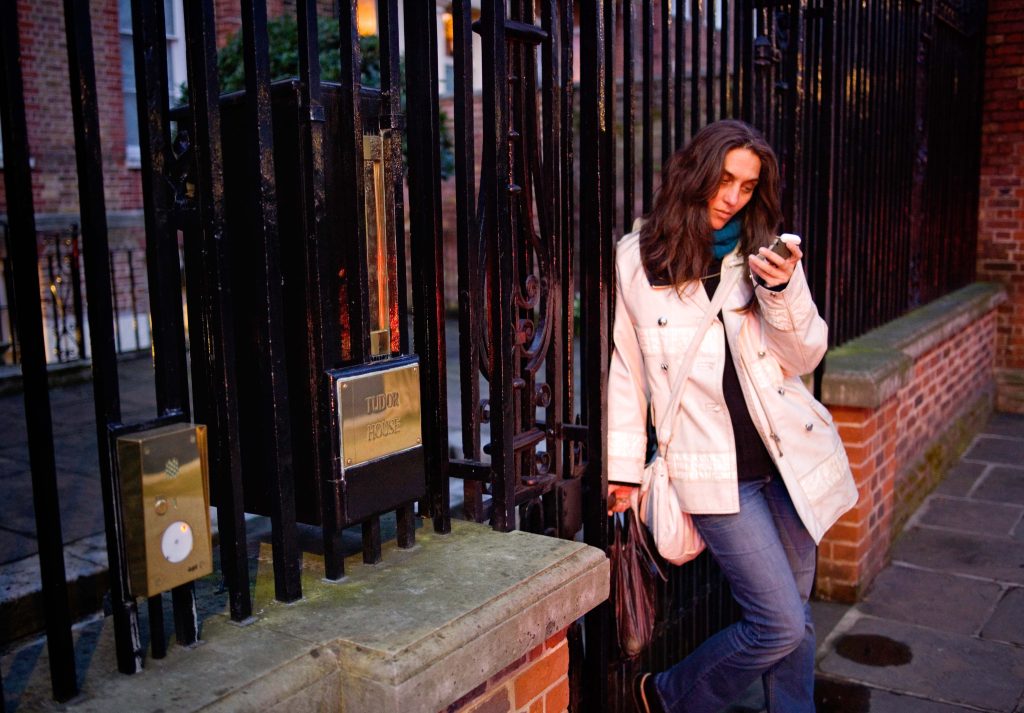 woman standing in front of rossetti's home at Cheyne Walk, London. Looking at her phone, a grocery bag in her hand, leaning against the gate.